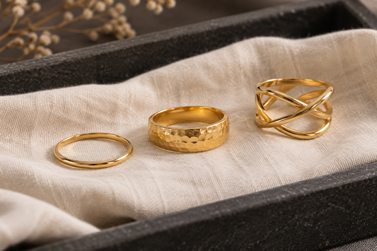 Three generic yellow-gold rings with slim, hammered and open crisscross profiles on ivory cloth.