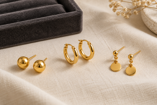 Three generic yellow-gold earring styles arranged on ivory cloth: ball studs, small hoops and dangling earrings.