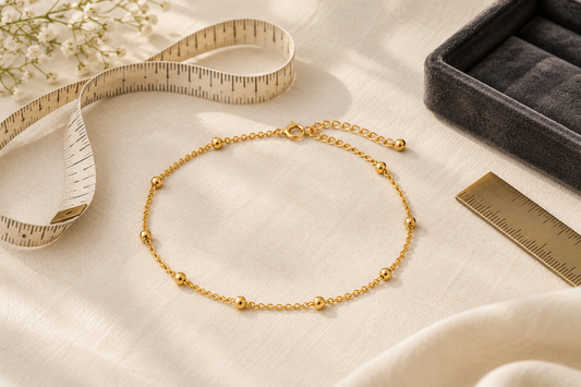 Generic yellow-gold anklet beside a soft measuring tape and jeweller's ruler on ivory fabric.