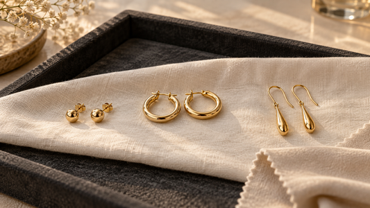Generic yellow-gold stud, hoop and drop earrings arranged on ivory cloth over a charcoal jeweller’s tray.