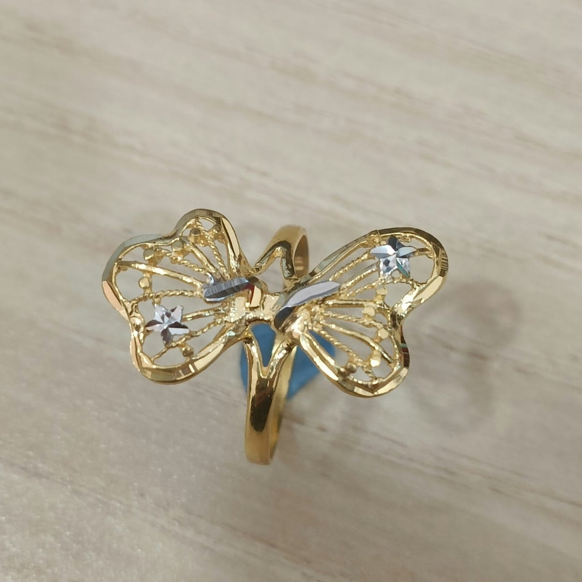 Front view of a 916 gold two-tone butterfly ring with openwork wings and silver-coloured details on pale wood