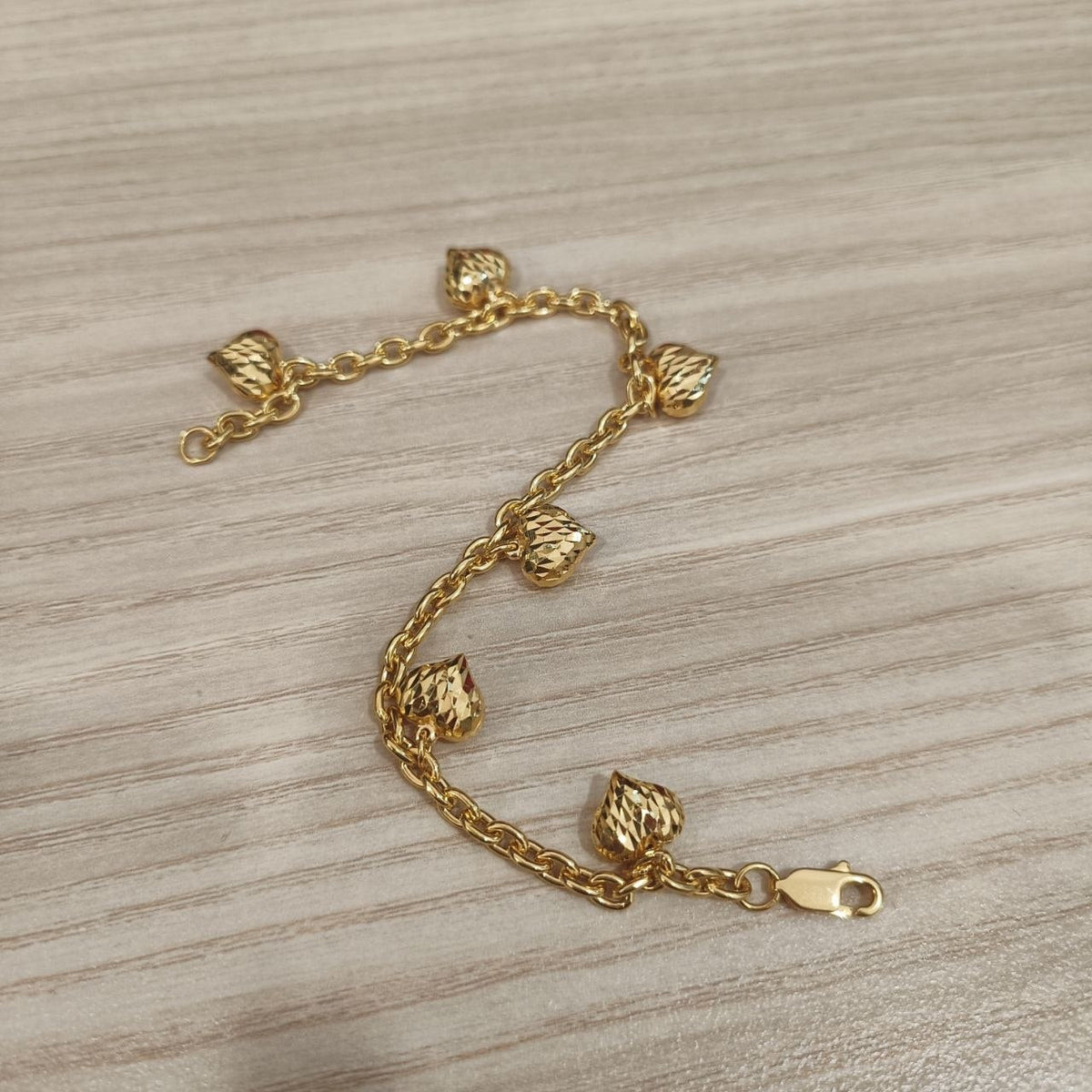 916 gold Wanzi six-heart bracelet arranged in an S curve showing faceted charms and clasp on pale wood