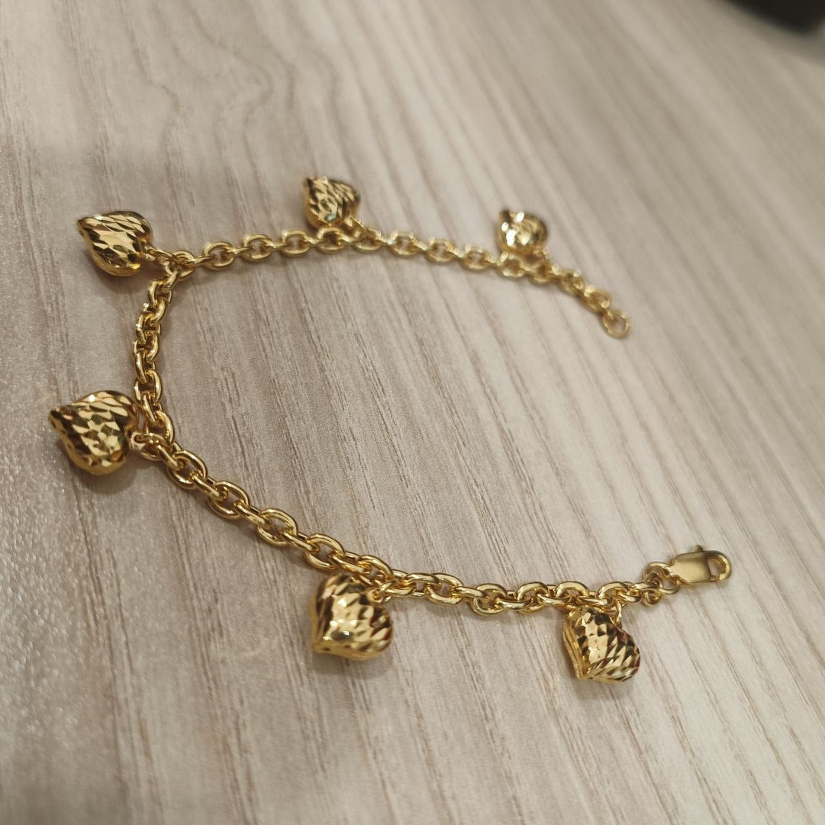 916 gold Wanzi bracelet arranged in an open curve showing textured heart charms and clasp on pale wood