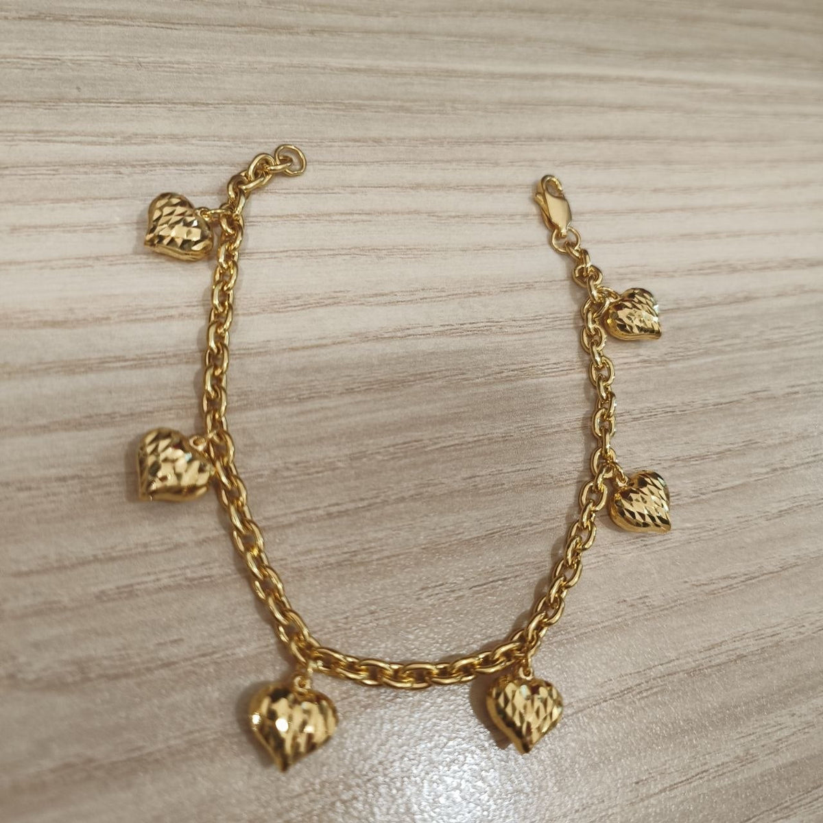 916 gold Wanzi six-heart bracelet laid in a U shape showing its chain, charms and clasp on pale wood