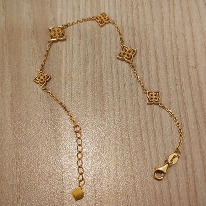 A gold chain bracelet with five small square quatrefoil motifs and an adjustable extender chain with a lobster clasp, displayed on a light wood grain surface.